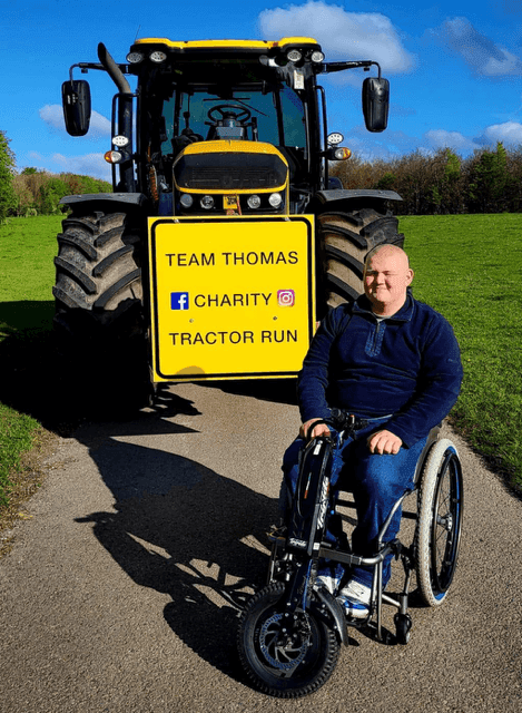Thomas in wheelchair in front of team sign on a tractor